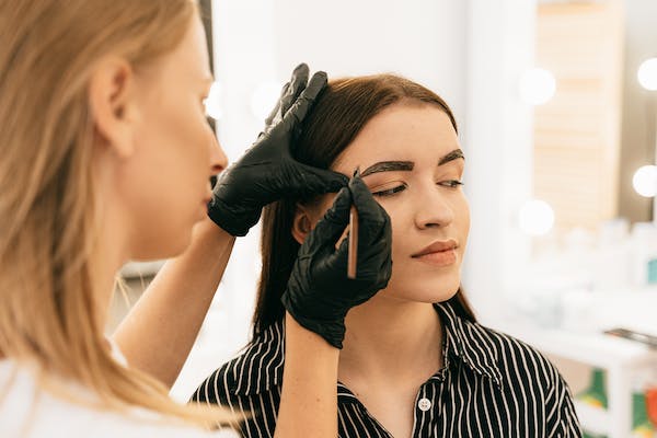 Female beautician shaping eyebrow of another seated woman.